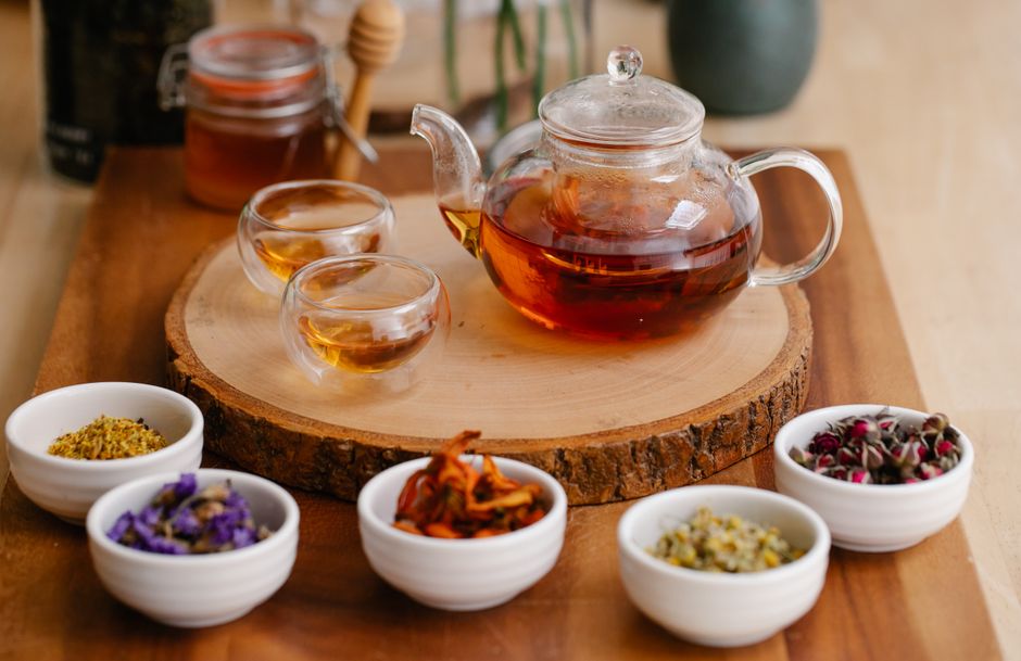 Glass teapot and cups with herbal infusions, fresh herbs, and honey dippers on a wooden tray.